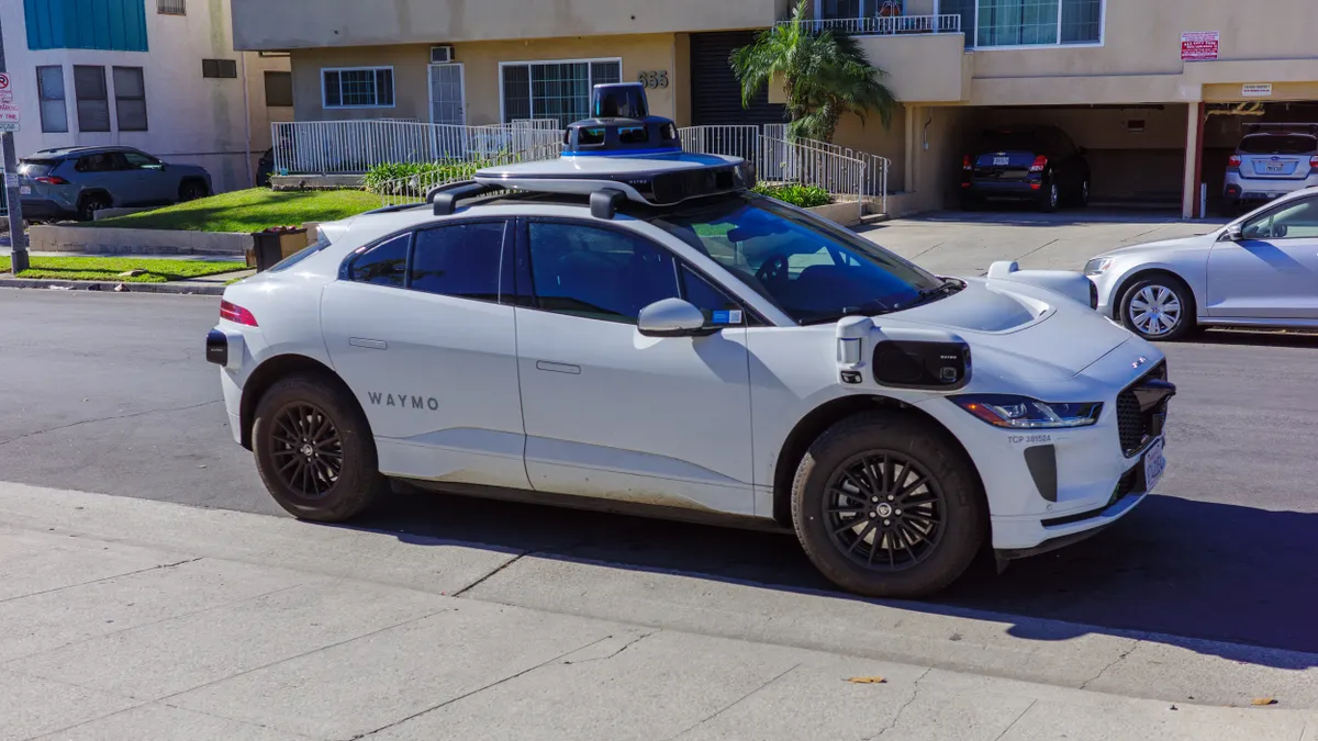 A white vehicle with sensors on the roof and sides of the car and a sign reading "Waymo" next to a sidewalk with apartment buildings across the street.