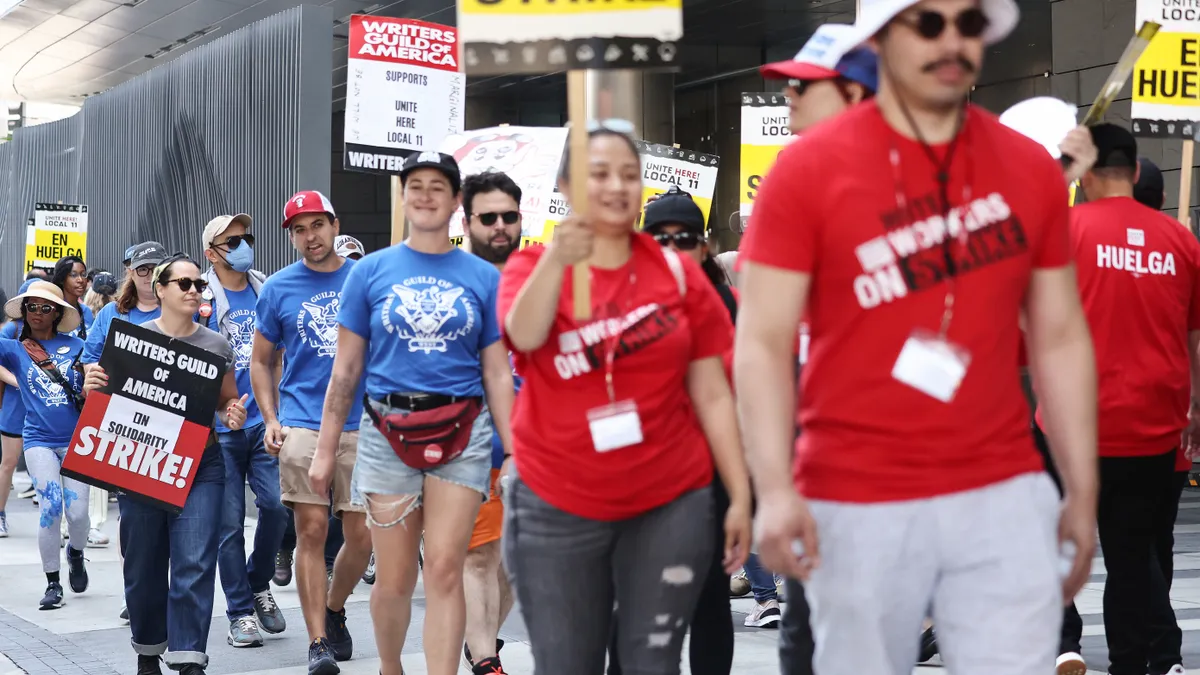 Workers in red Unite Here shirts hold picket signs on a sidewalk.