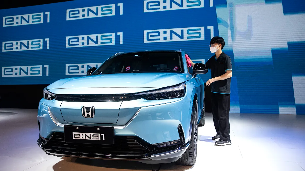 A man stands with a blue Honda in the foreground, on display. He wipes the driver's side window with a pink cloth.