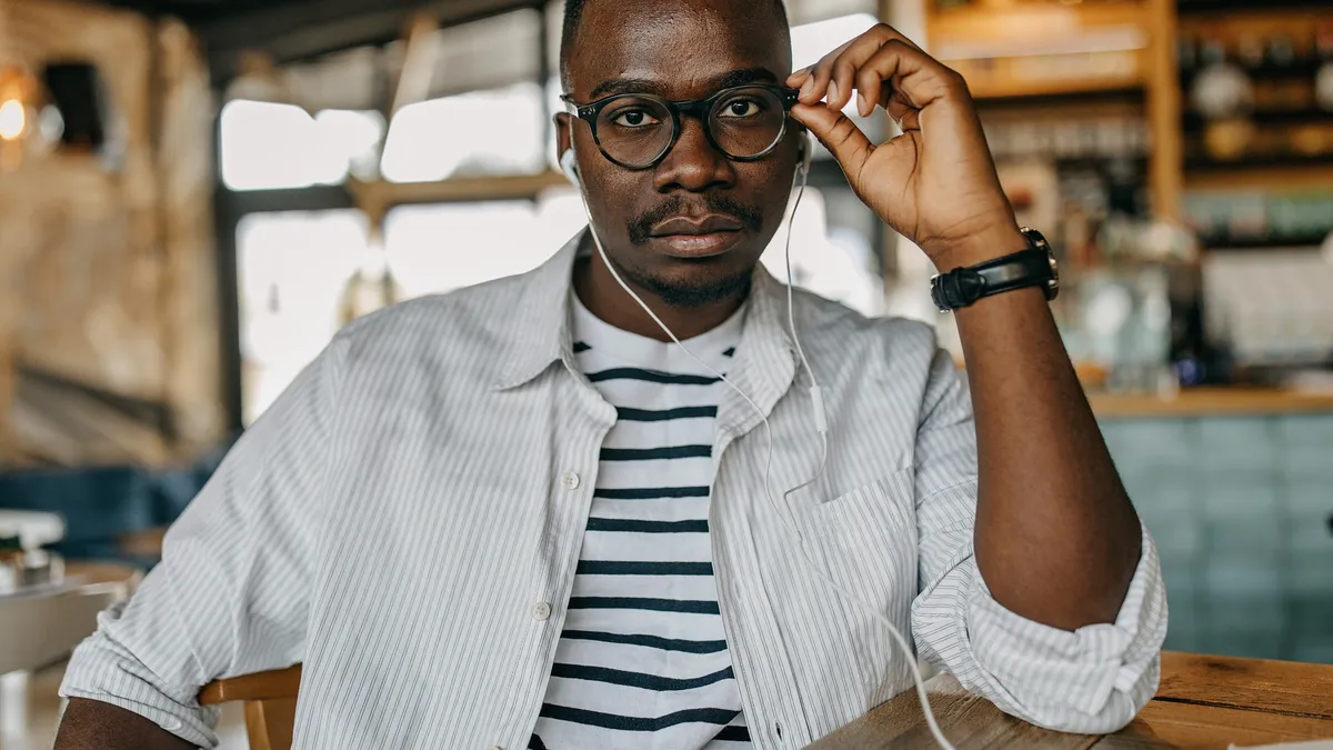 A portrait of a pensive young Black man with glasses and a hightop fade in a coffee shop