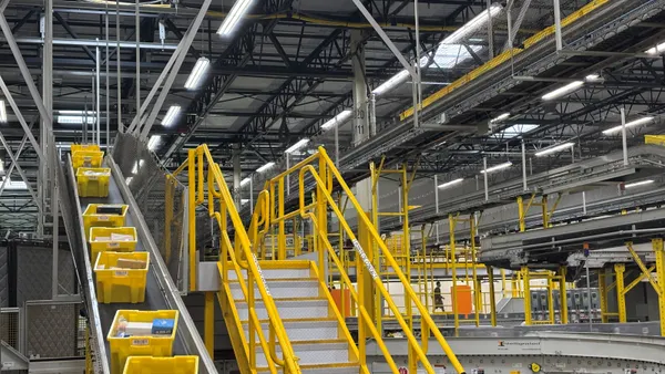 Yellow bins move on a conveyor belt located next to a set of stairs with yellow railings within a warehouse.