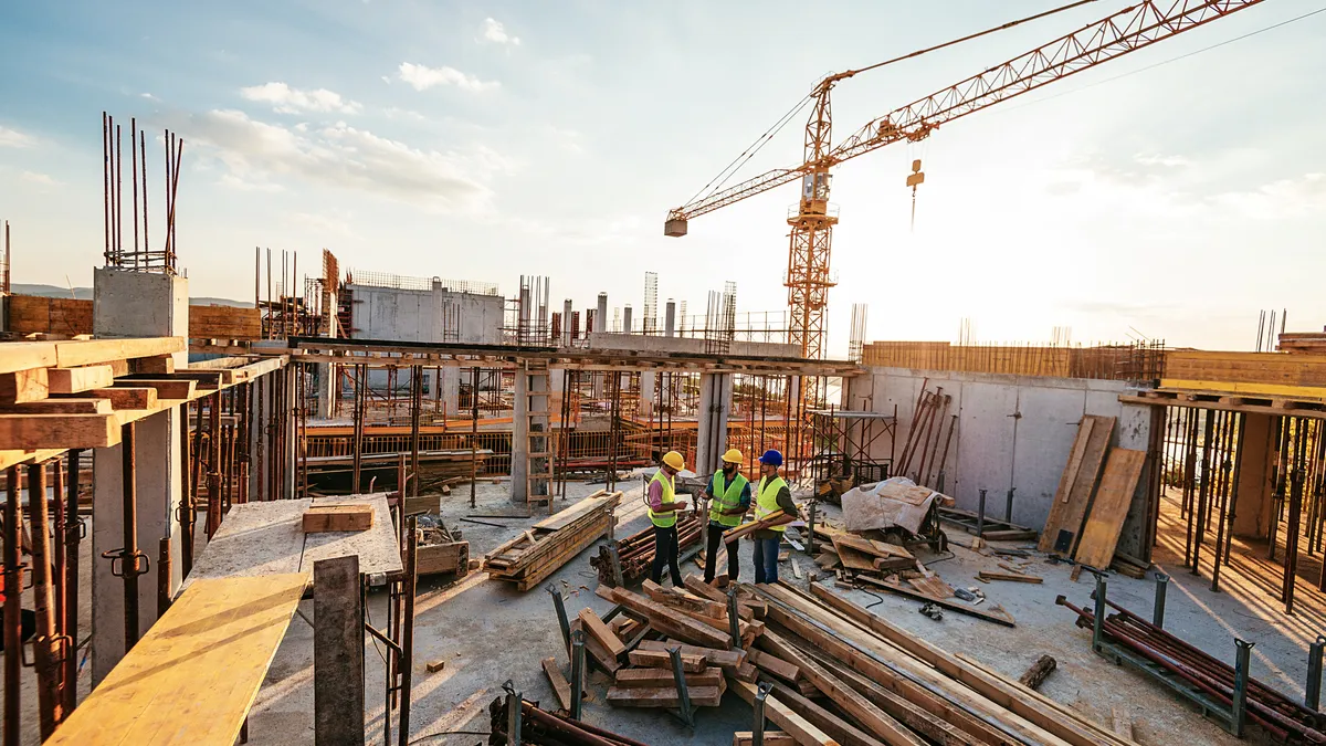 Three construction workers are in the middle background, surrounded by work on a large construction site.