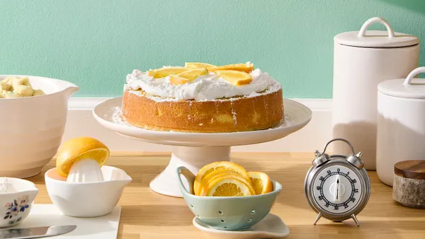 A cake on a white cake stand against a mint green wall. A bowl of lemon slices is positioned in front.