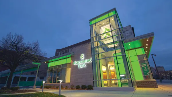 A glass and stone building is shown in the evening, with lights on inside, and "Associated Bank" name and logo on the outside.