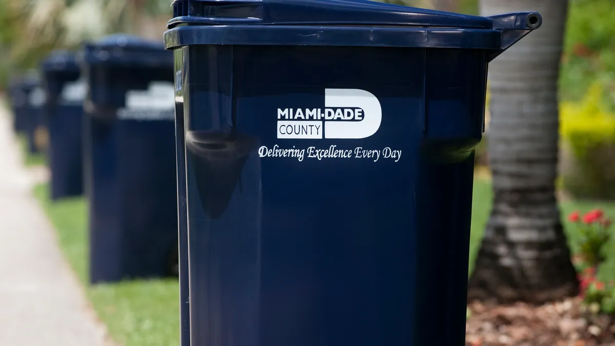 A line of trash bins stamped with "Miami-Dade County, Delivering Excellence Every Day"