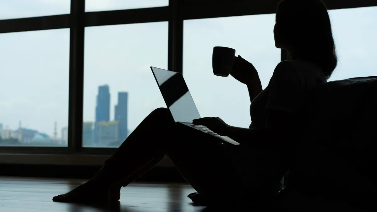 The silhouette of an individual working on a laptop in front of a window.