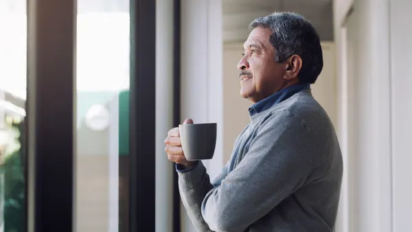 Man smiles while holding a mug and looking out the window.