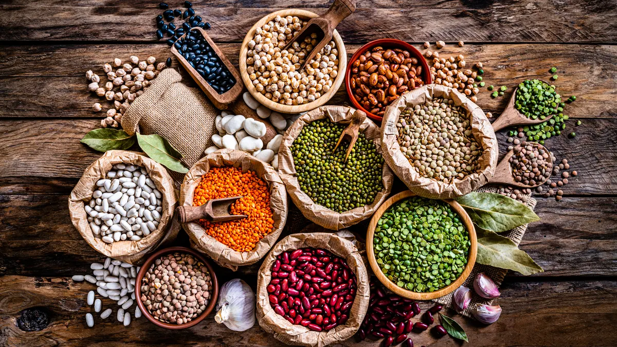 Overhead view of several sacks and bowls of various plant-based ingredients and legumes, some with scoops, grouped together on a wooden surface.