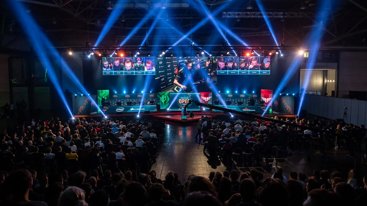 An esports stage light up with blue lighting is watched by a crowd.