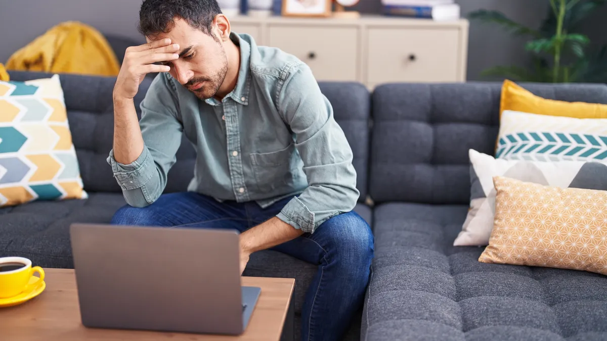 A stressed man uses his laptop while sitting on a couch at home.