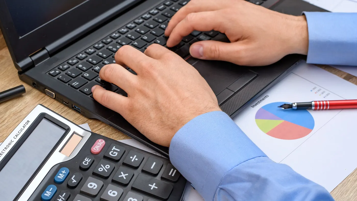 A man working at a desk, typing on a computer, a calculator and a pie chart lying next to it, closeup.