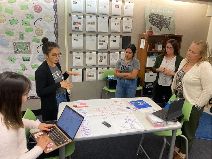 Five adults are standing around a table inside a classroom. Papers are on the table and one person is holding a laptop computer.