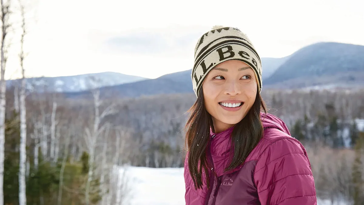 A person wearing a knit L.L. Bean hat looks off to the right, with trees and mountains in the background.