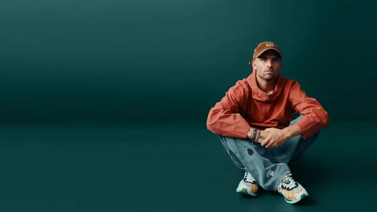 person wearing a brown cap, orange jacket, jeans and sneakers sits cross-legged, off to the right, against a blue-green background.