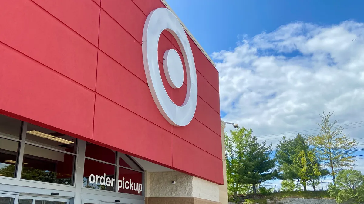 A red storefront entrance with a white dot-in-circle logo and an "order pickup" sign on the door. Trees and blue sky with white clouds on the right.