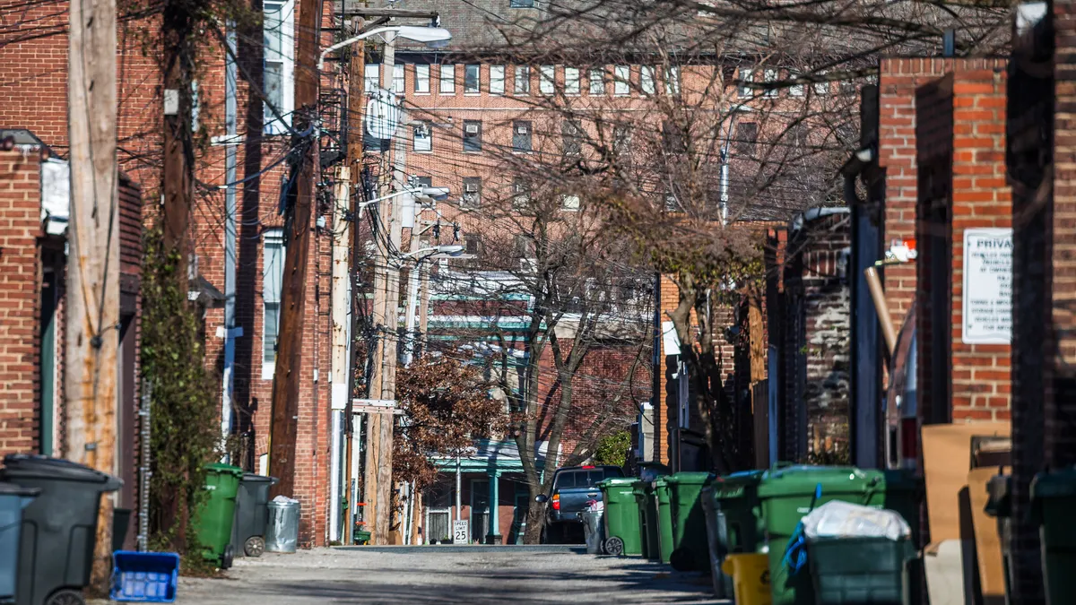 View of a back alley lined with trash carts in Baltimore, with utility lines and tree branches overhead