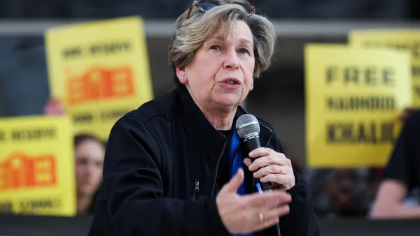 A person speaks using a microphone as they stand in front of a crowd holding yellow protest signs.
