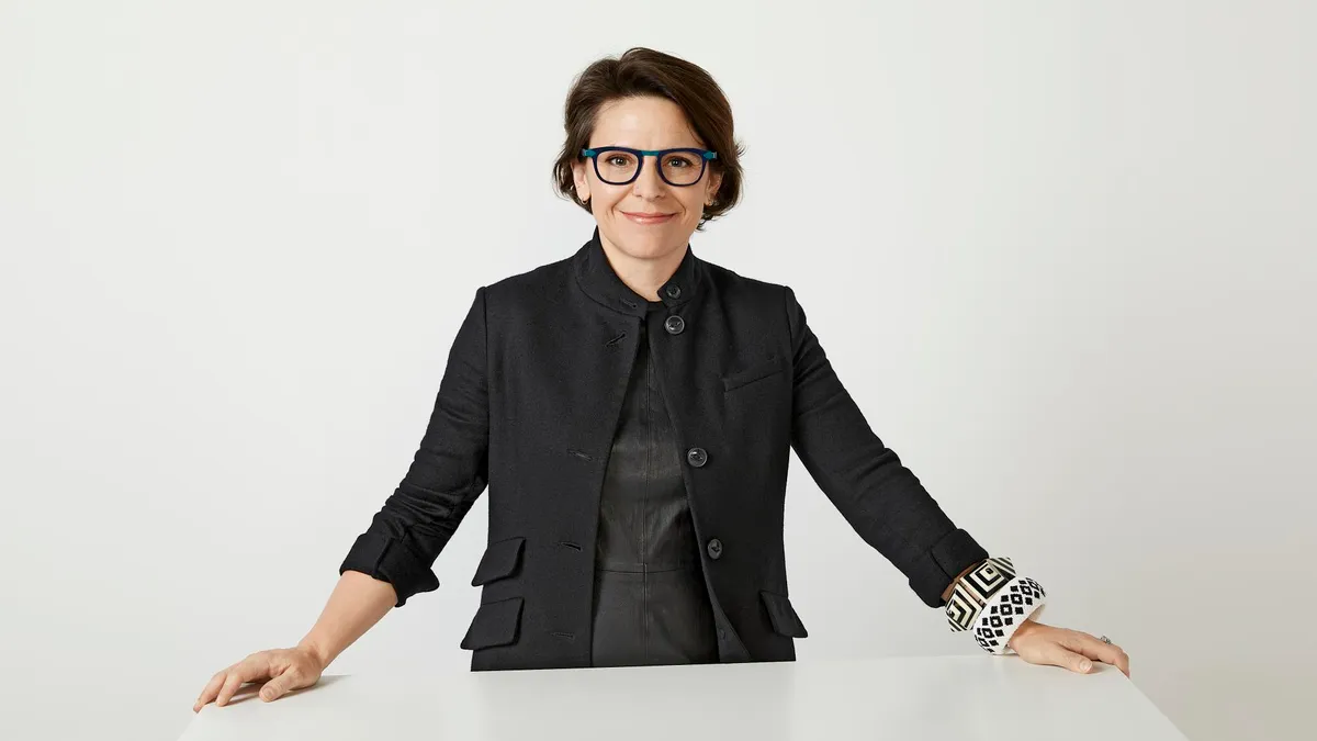 A dark-haired person wearing black-rimmed glasses, black clothes and black-and-white wrist cuffs smiles slightly against a white background.