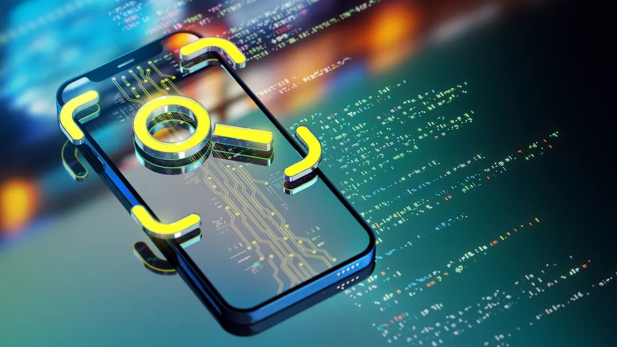 A yellow loupe icon seen on top of a smart phone and data code seen on a monitor behind it.