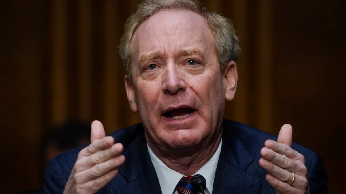 A close up of a man in a blue suit with a multicolored tie gesturing while seated at a desk.