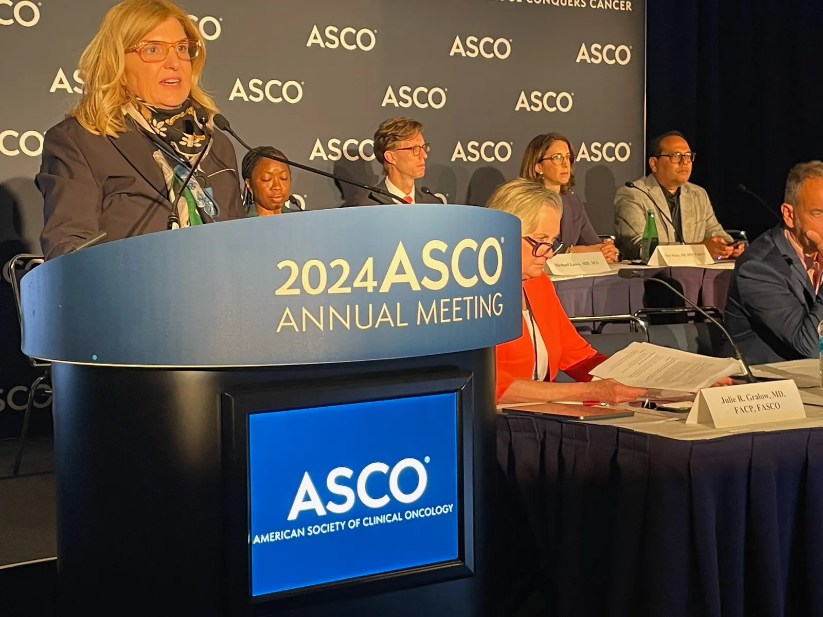 A person speaks at a lectern next to several others seated at a lower table.