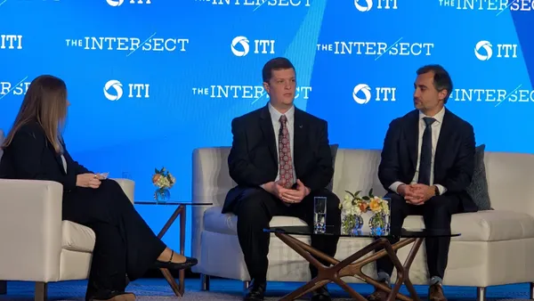 Two men and a woman sit on an event stage in front of a blue backdrop that displays the name of the event and its organizer