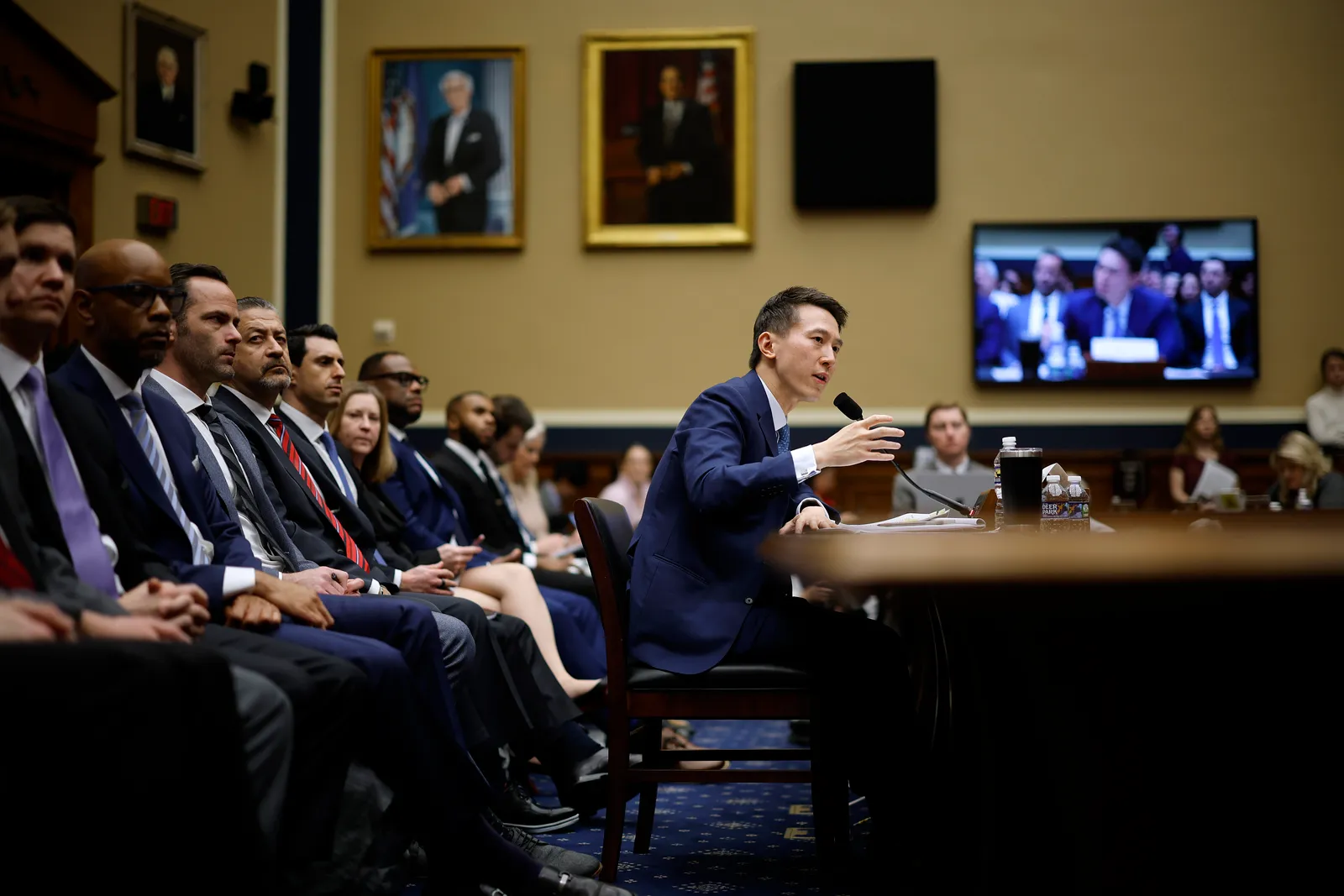 An adult sit at a desk and appears to be speaking into a microphone. The adult is in a large hearing room and there are rows of people seated behind the person. The camera angle is from the side.