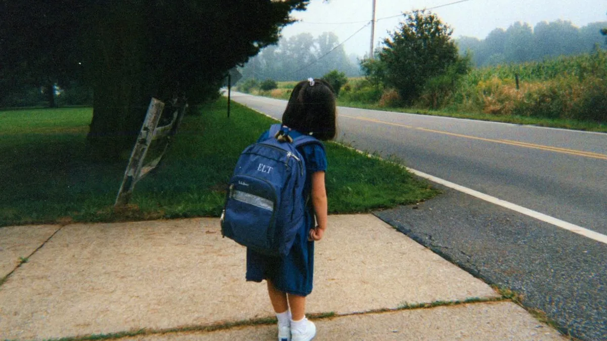 A child in a blue dress stands at the end of a driveway waiting for the bus to arrive while wearing a blue L.L.Bean backpack.