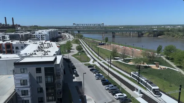 Aerial view of a tram alongside a river with buildings on the left and a bridge over the river in the distance.