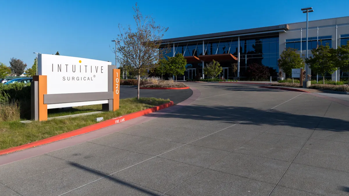 A modern office building with large reflective glass windows is shown behind a wide driveway and landscaped entrance. In the foreground, a white and orange monument sign reads "Intuitive Surgical" with the number "1020" vertically on the side.