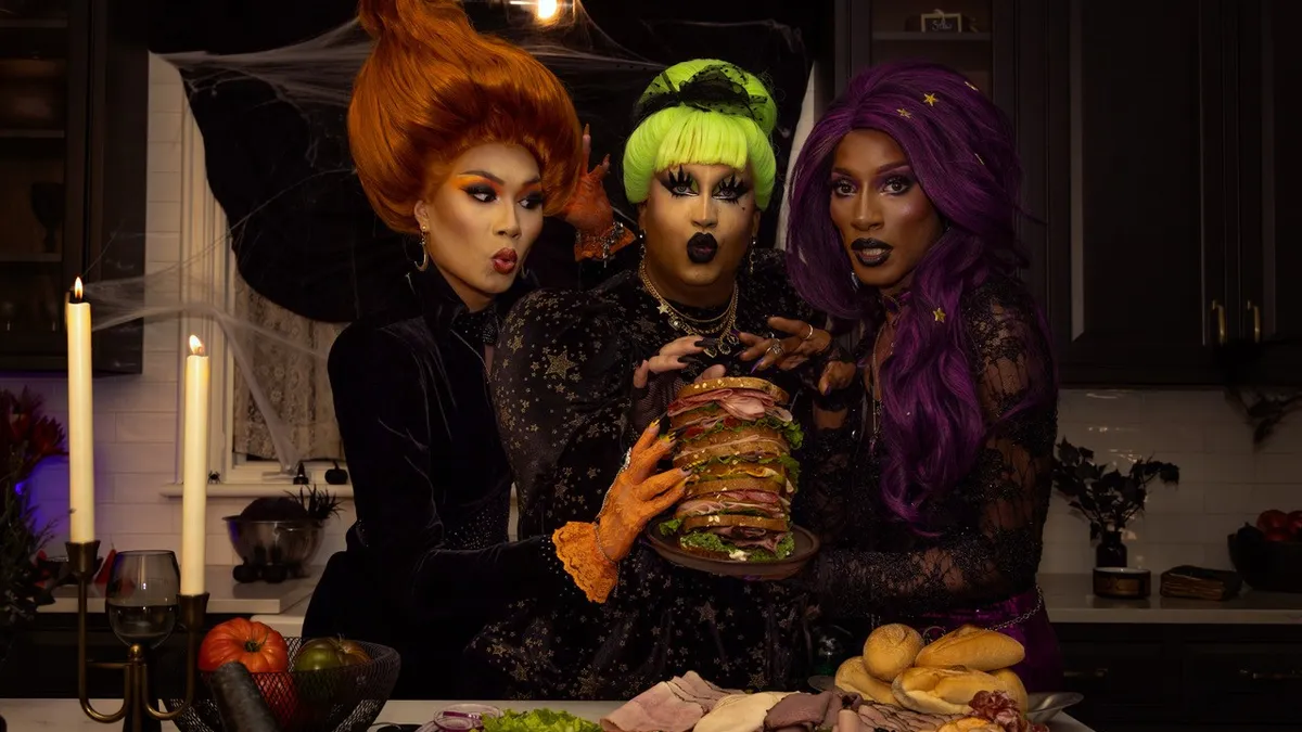 A trio of drag stars dressed as witches hold a stacked sandwich over a kitchen table for a new Dietz & Watson Halloween campaign.