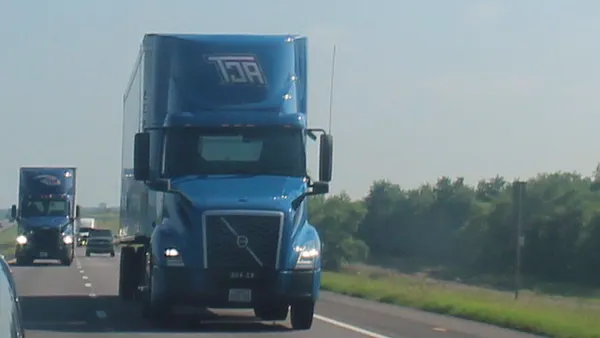 A passenger vehicle mirror shows the front of an ACT tractor driving along a highway.