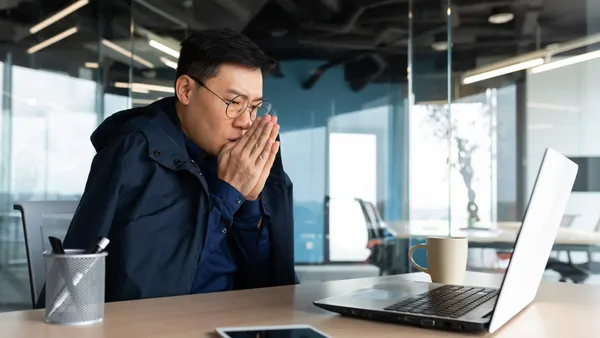 An office worker is bundled up and cold, looking at a laptop.