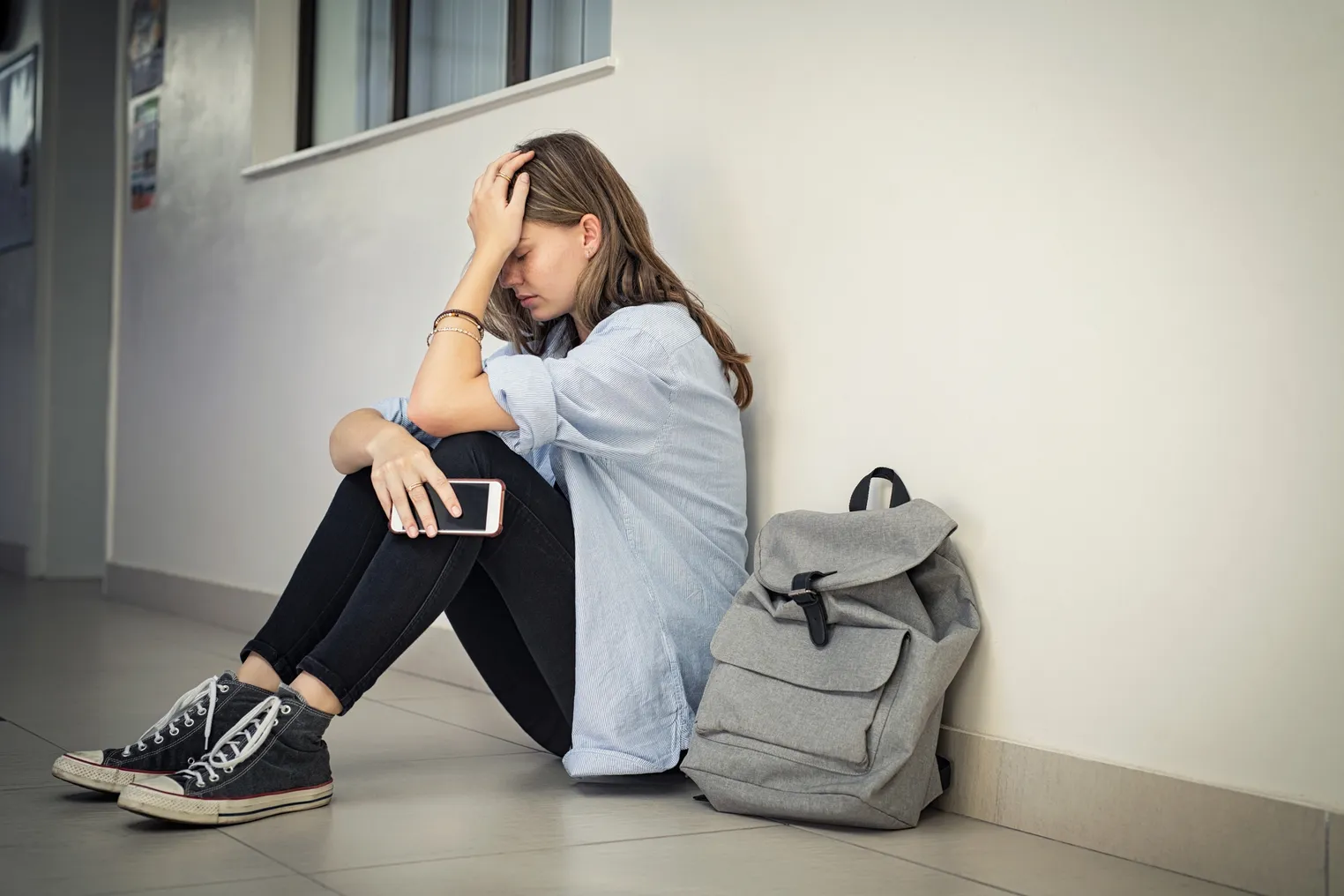 A high school student sits in a school hallway with head in hand and cell phone in other hand.