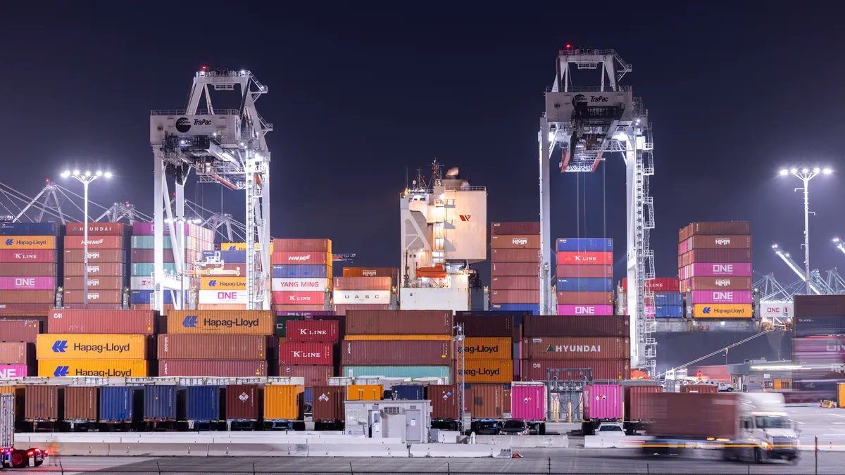 Cranes unload intermodal containers from a ship at the Port of Los Angeles at night, with a truck in motion in the foreground.
