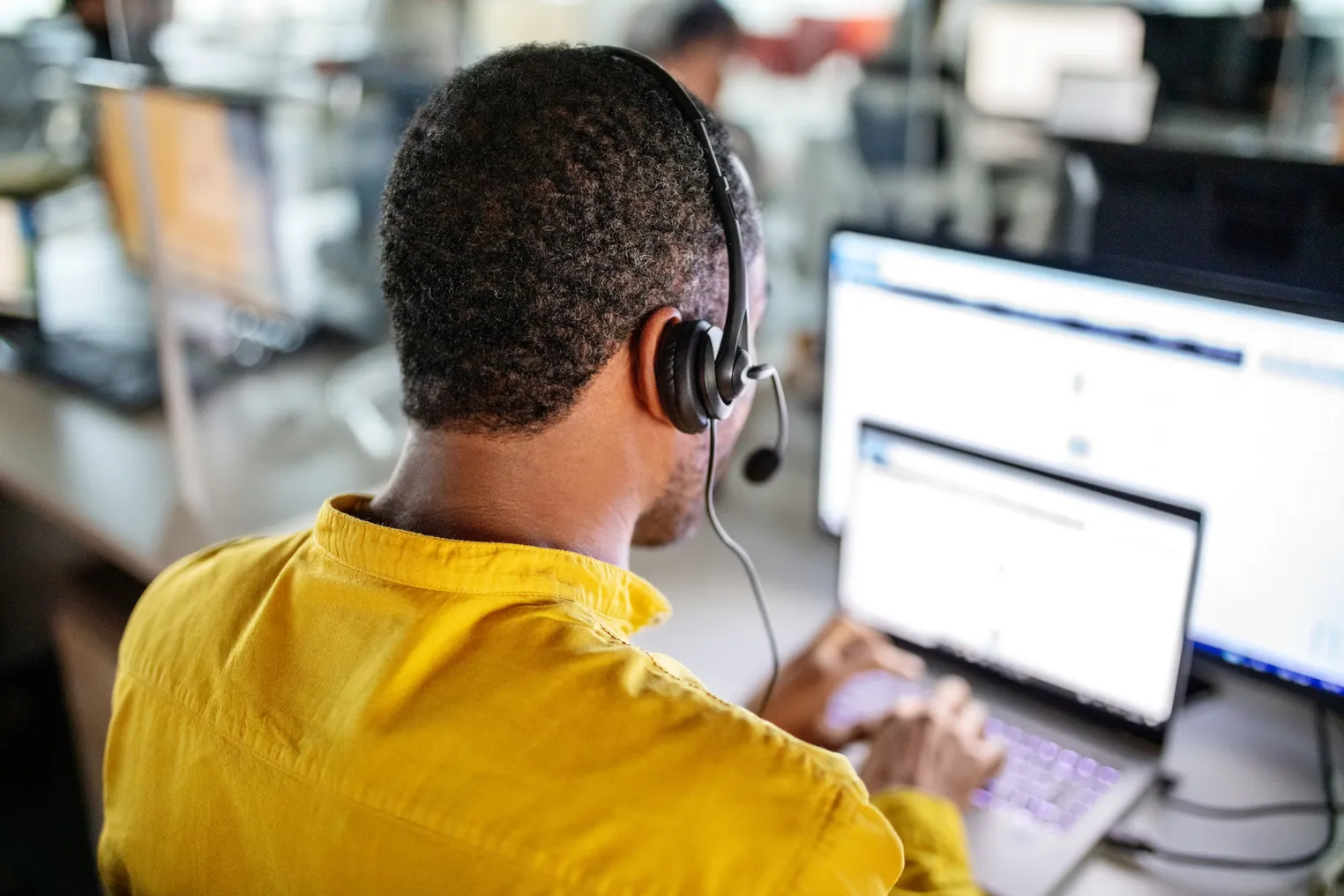 A customer service or call center employee sits at a desk while using a laptop, headset with microphone and monitor.