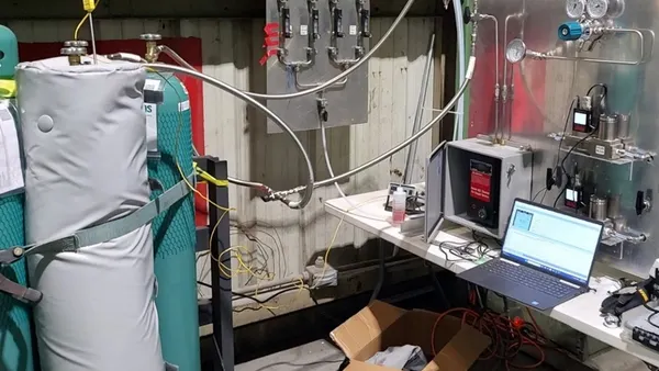 A complex setup showing pressurized and insulated tall tanks hooked up to pipine system with a stainless steel wall of devices. A laptop also sits on a table hooked up to the instruments with wires.
