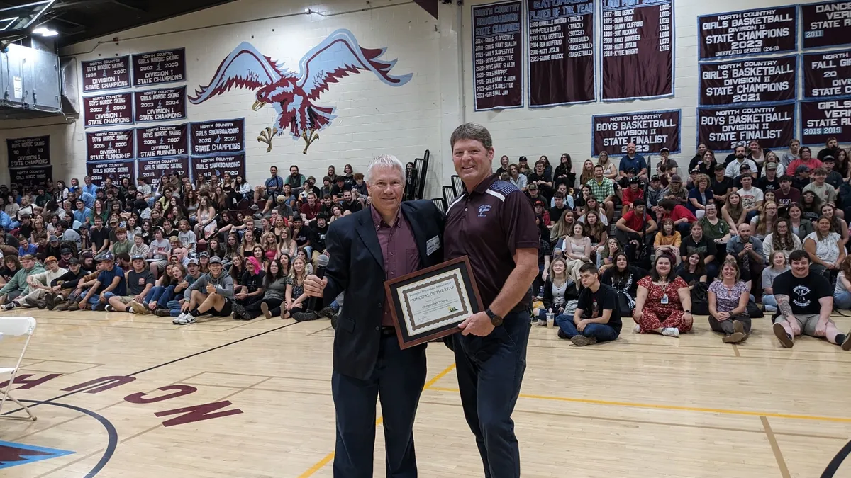 Two adults stand on a basketball court in a school gym holding a certificate. In the background are bleachers filled with people.