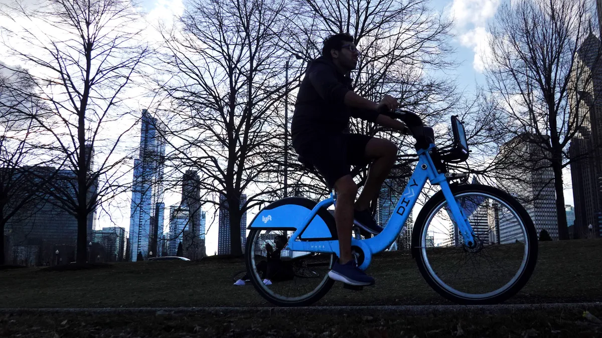 Person riding a blue Lyft bike against a background of barren trees and tall buildings.