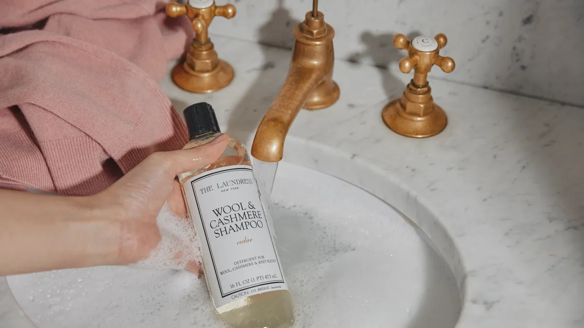 A hand holding a wool and cashmere shampoo bottle from The Laundress over a marble sink