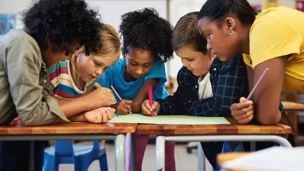 Five students are sitting close together at a table. They have pens and markers in their hands and they are writing on a sheet of paper on the desk.