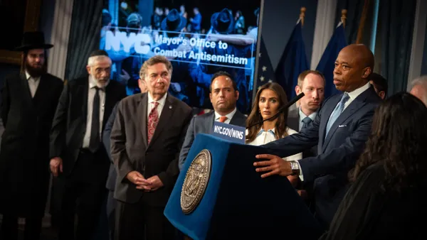 A person speaks at a podium surrounded by other people in front of a sign with the words "NYC Mayors Office to Combat Antisemitism."