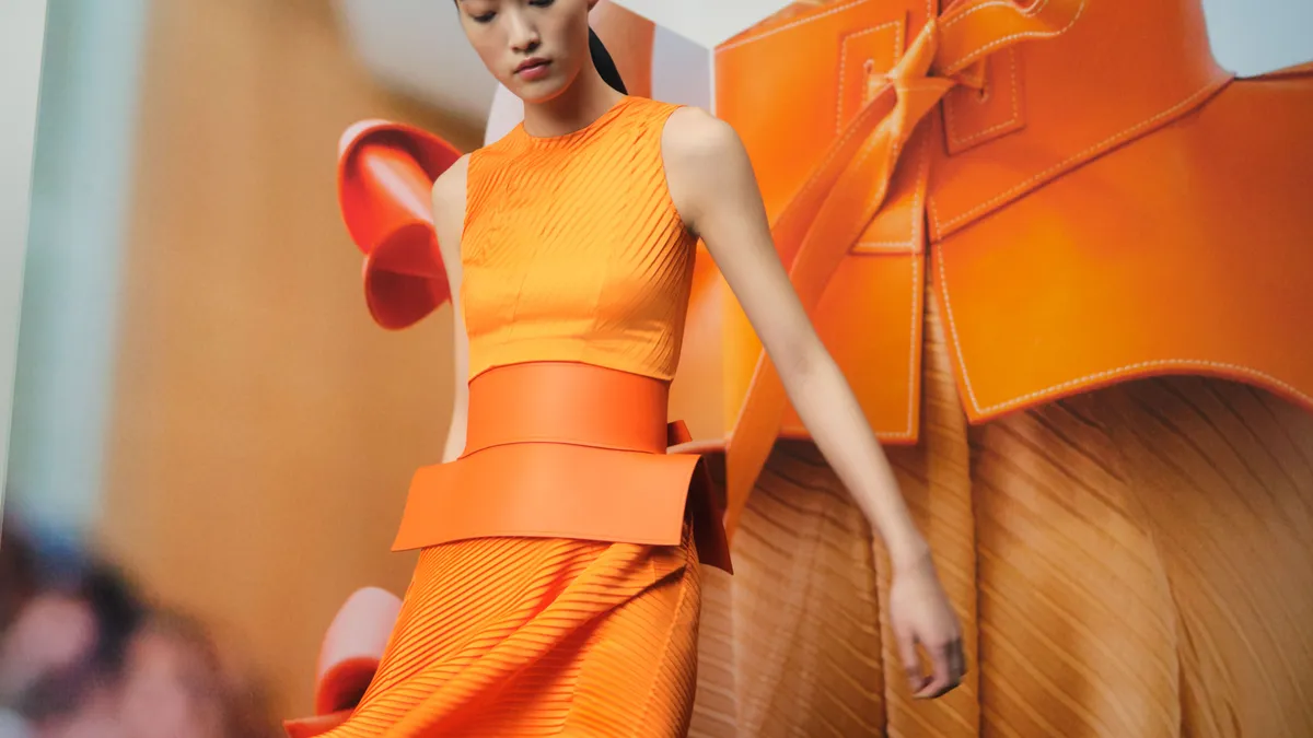 A model dressed in orange wears a wide orange belt and stands in front of a photo of the belt.