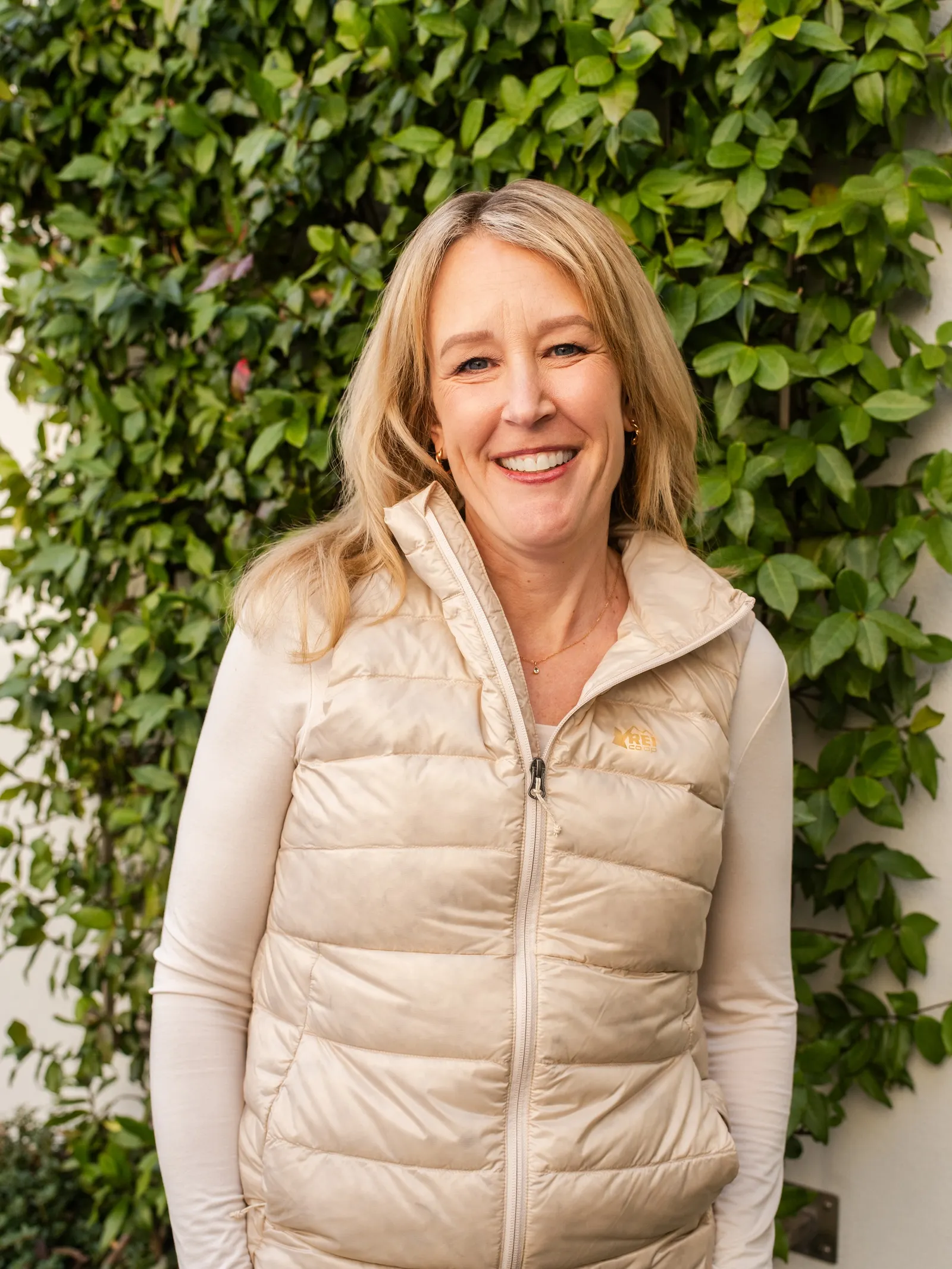 A person in a cream-colored vest stands in front of a wall of foliage.