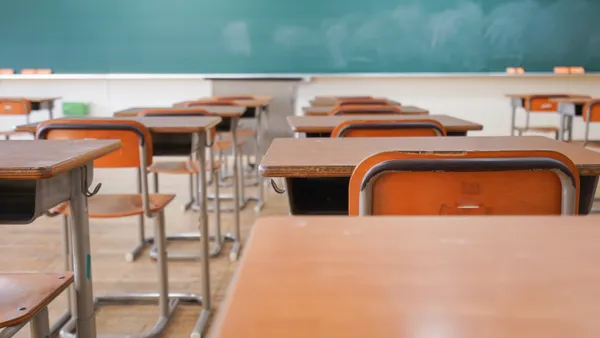 Rows of empty chairs and desks sit in front of a blank chalkboard in a school classroom.