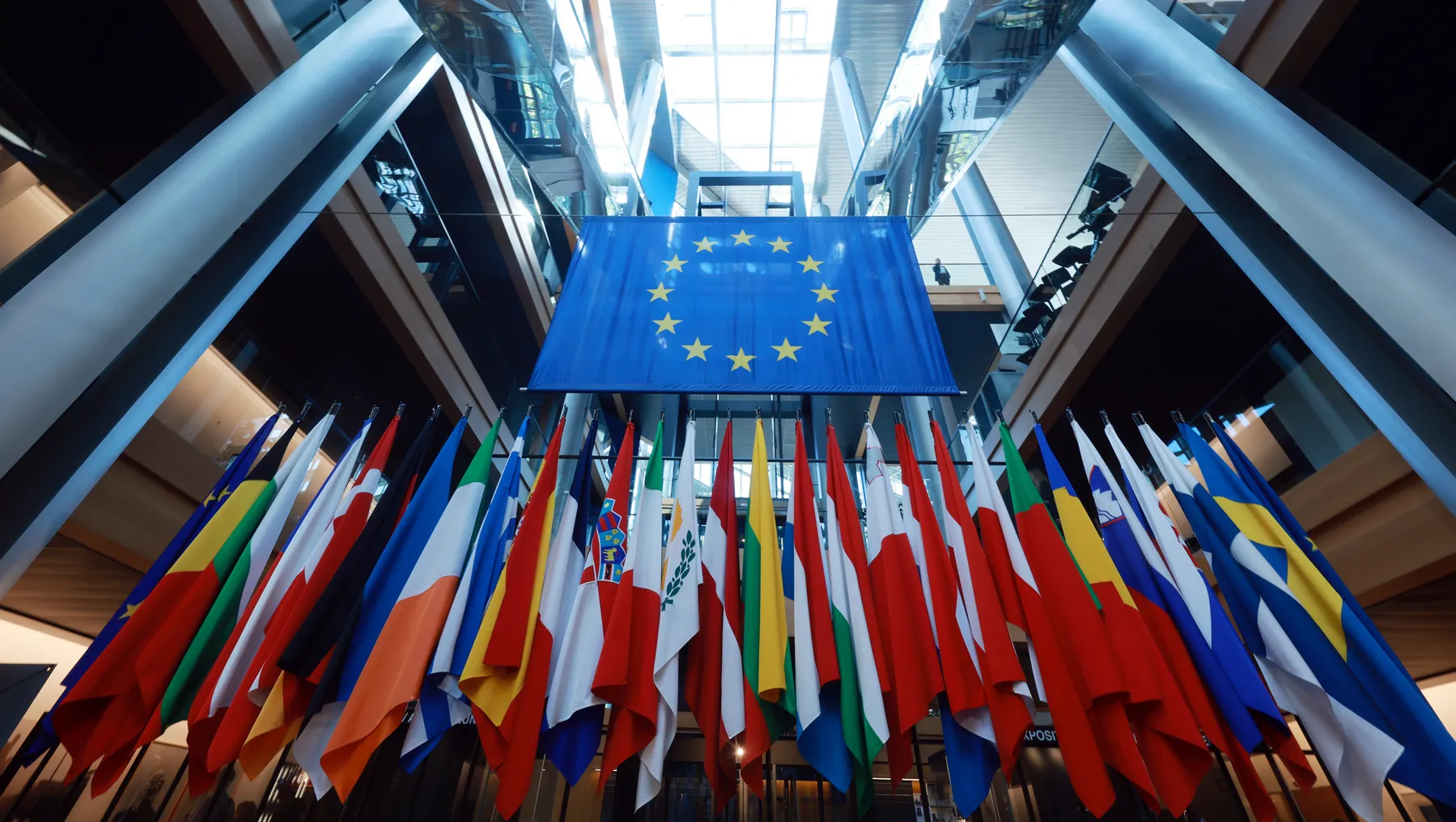 A group of flags on flag poles stand underneath one large blue flag, which features a circle of gold stars in the middle.