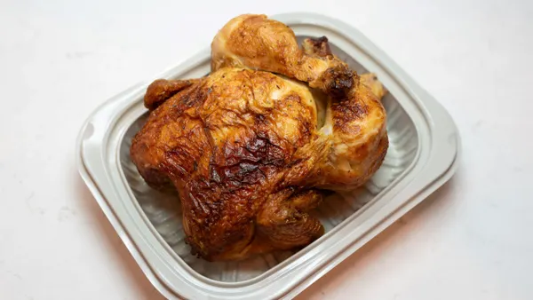 A full rotisserie chicken sits inside the bottom of a beige plastic clamshell container with the lid off.