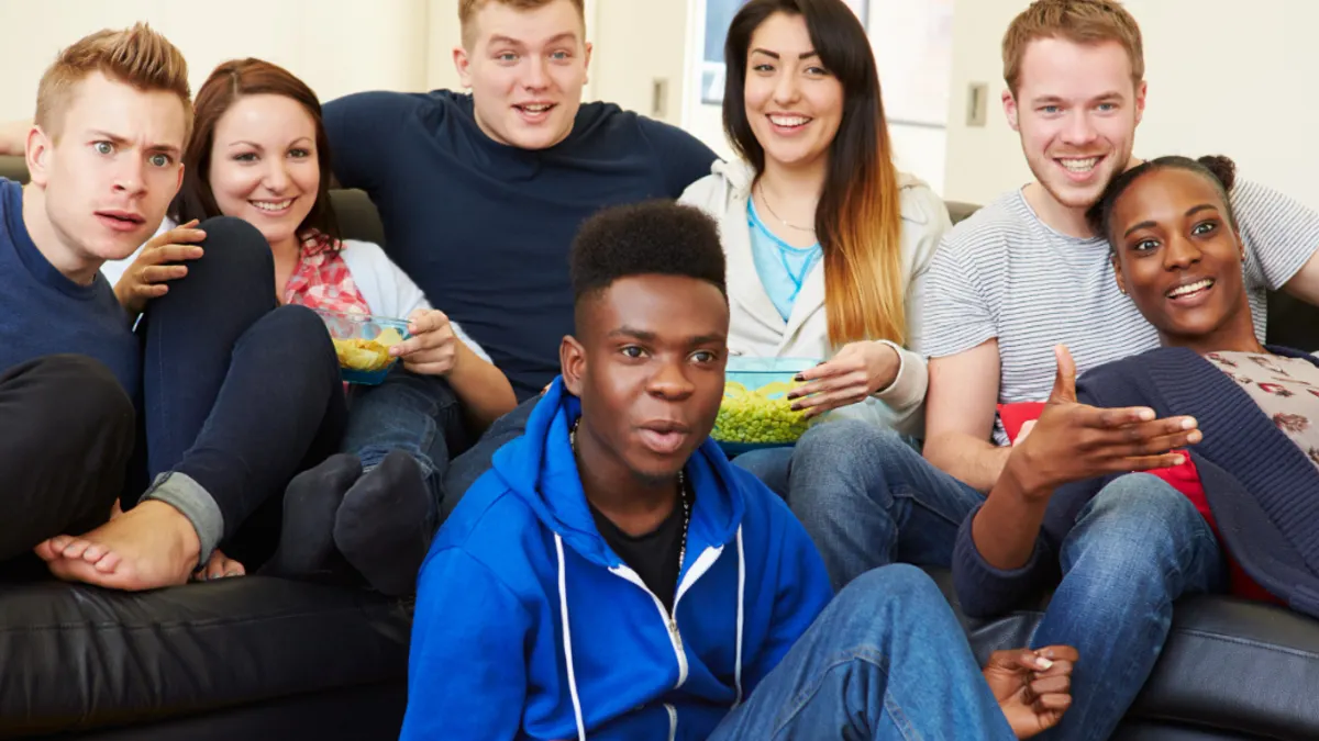 Six friends sit on a couch watching TV with snacks.