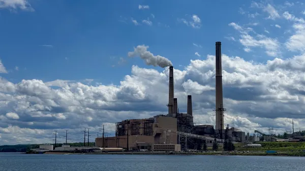 Concrete buildings with smoke stacks emitting white smoke into blue sky with body of water in foreground.