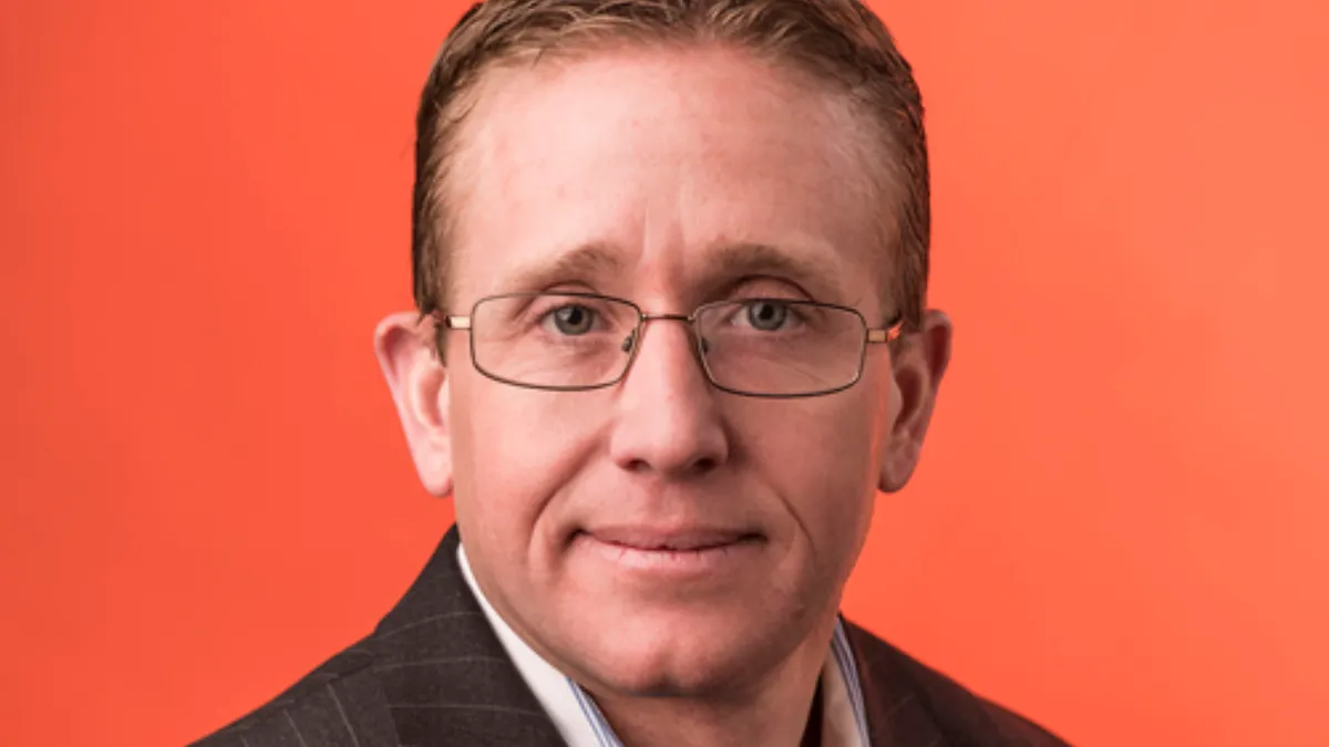 A headshot of a person with eye glasses and a brown business coat with a red background.